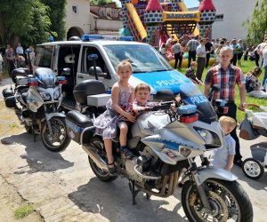 Zdjęcie przedstawia dwójkę dzieci siedzących na motocyklu policyjnym, drugi motocykl policyjny, radiowóz oraz innych uczestników dnia dziecka.