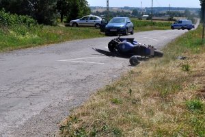 Zdjęcie przedstawia przewrócony motocykl, kask, stojące samochody na poboczu oraz radiowóz.
