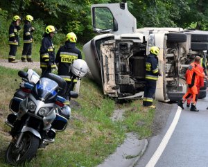 Zdjęcie przedstawia policyjny motocykl, przewrócony samochód ciężarowy i strażaków.
