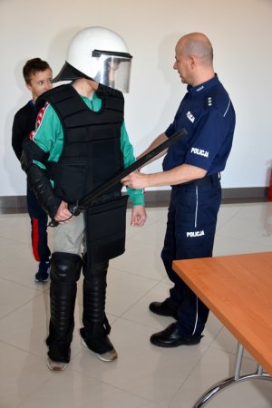 Zdjęcie przedstawia policjanta ubierającego ucznia w strój oddziałów prewencji.