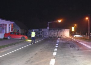 Zdjęcie przedstawia policjanta przy zdarzeniu drogowym, dwa samochody osobowe na poboczu oraz przewrócony samochód ciężarowy.